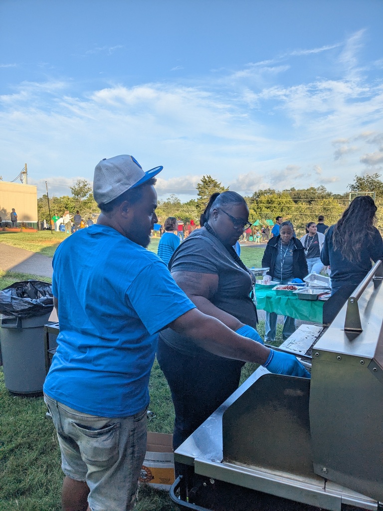 Two people in blue and black clothing prepare food on a grill. A table with food is behind them.