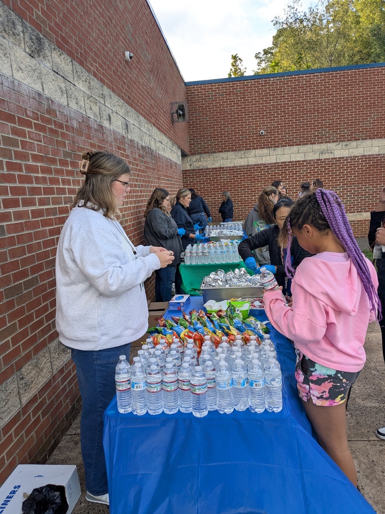 A group of people outside. One woman talks to a young girl. A table has many items and water bottles.