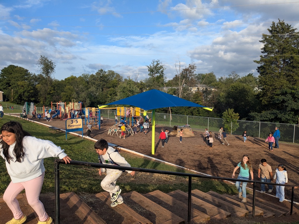 Children play and run at a playground. Blue canopy over sand pit. Some have backpacks. Trees and fence in background.