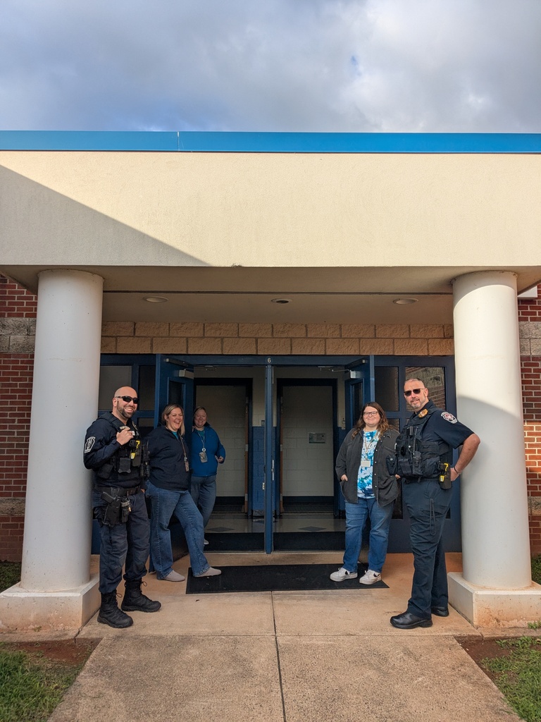 Police officers stand outside a building. Two officers have their hands on their hips, while three people stand in the open doorway.