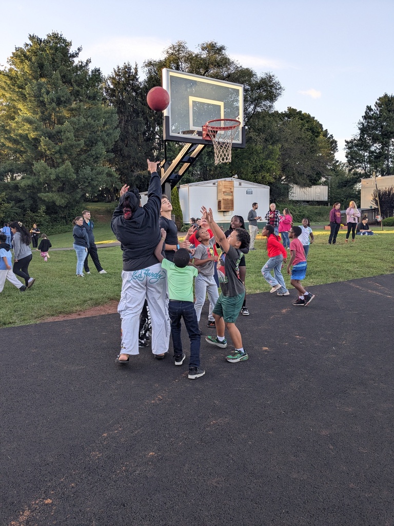 A group of people plays basketball outdoors on a paved court. Some reach for the ball. Green trees are in the background.