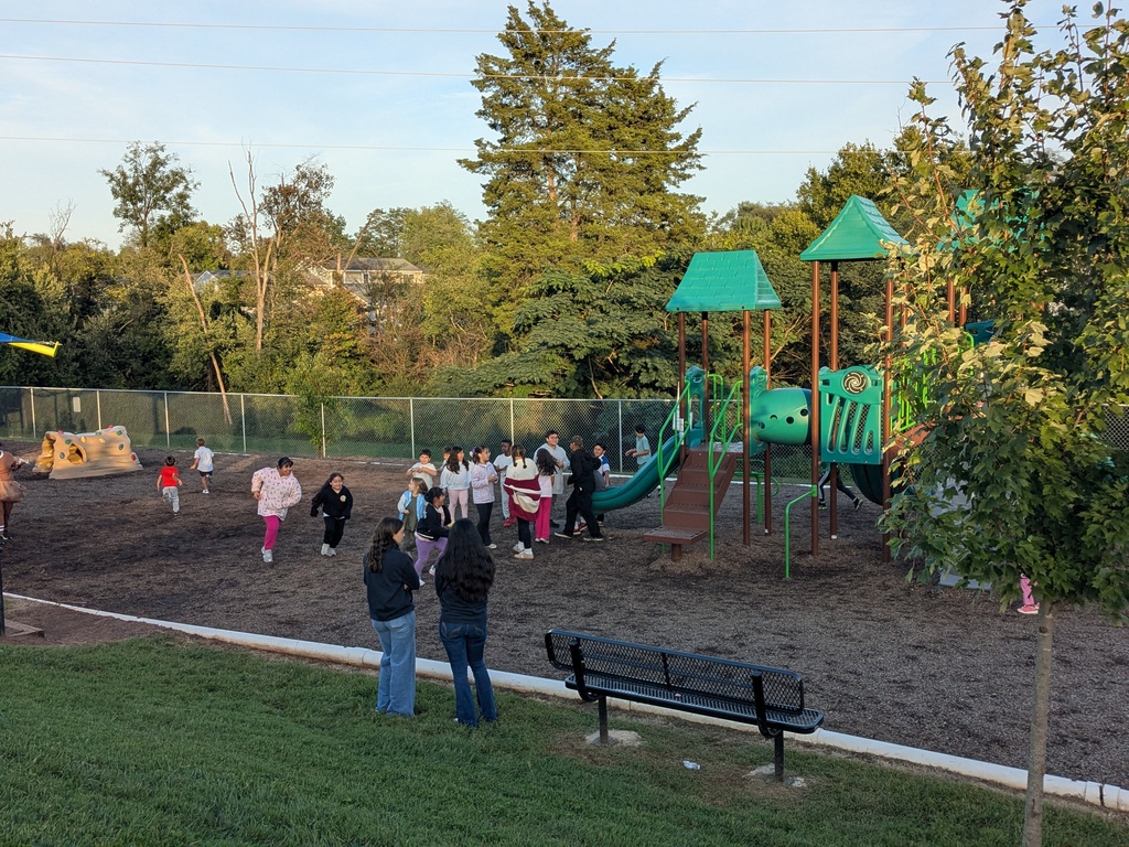 Several people, including children, stand or walk near a playground in a park. The playground has slides and climbing equipment. A bench and tree are in the foreground.