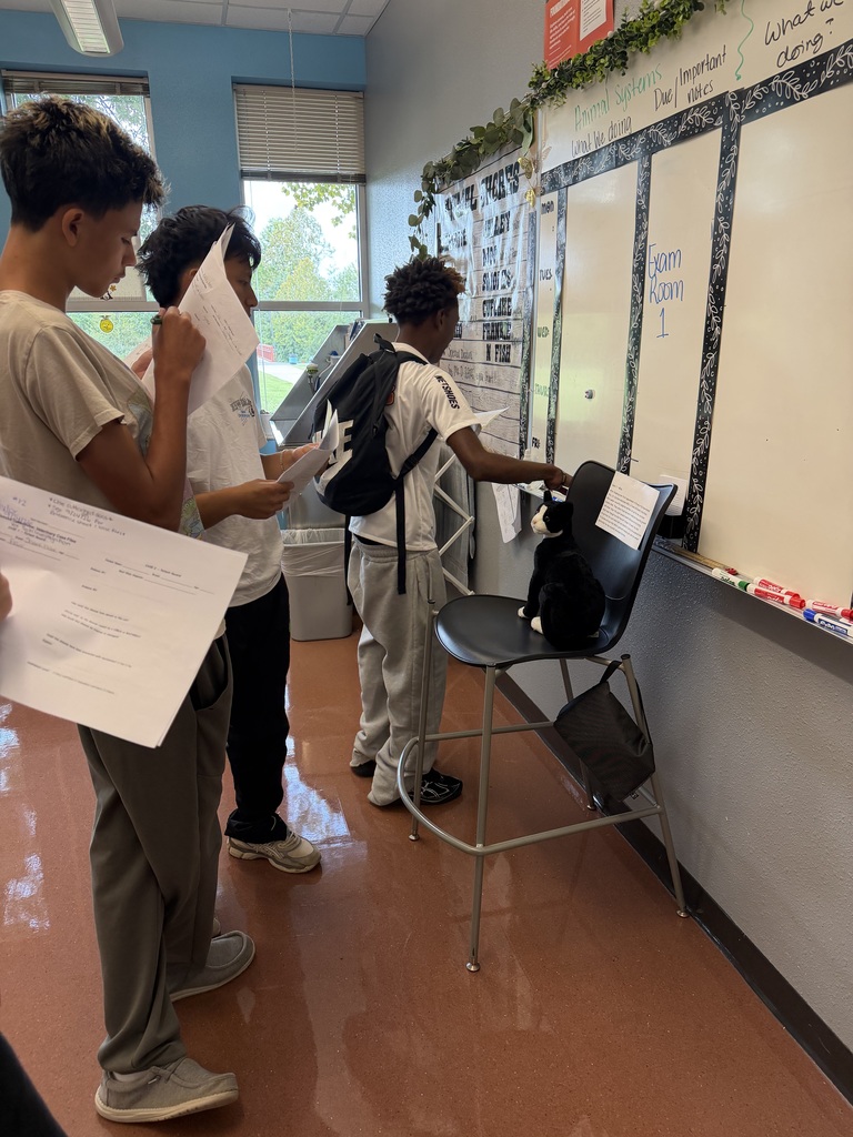 Three students stand in a room, holding papers and a backpack, with a chair and bulletin board nearby.
