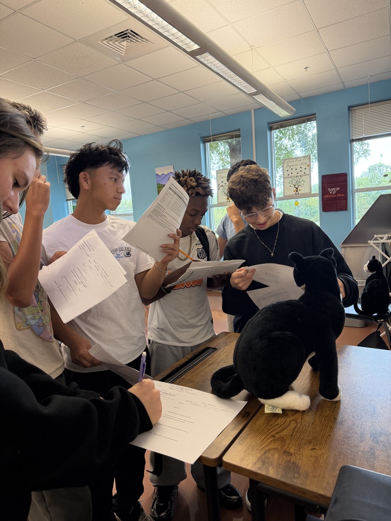 A group of students stands around a table in a room. They are holding papers and a stuffed animal.