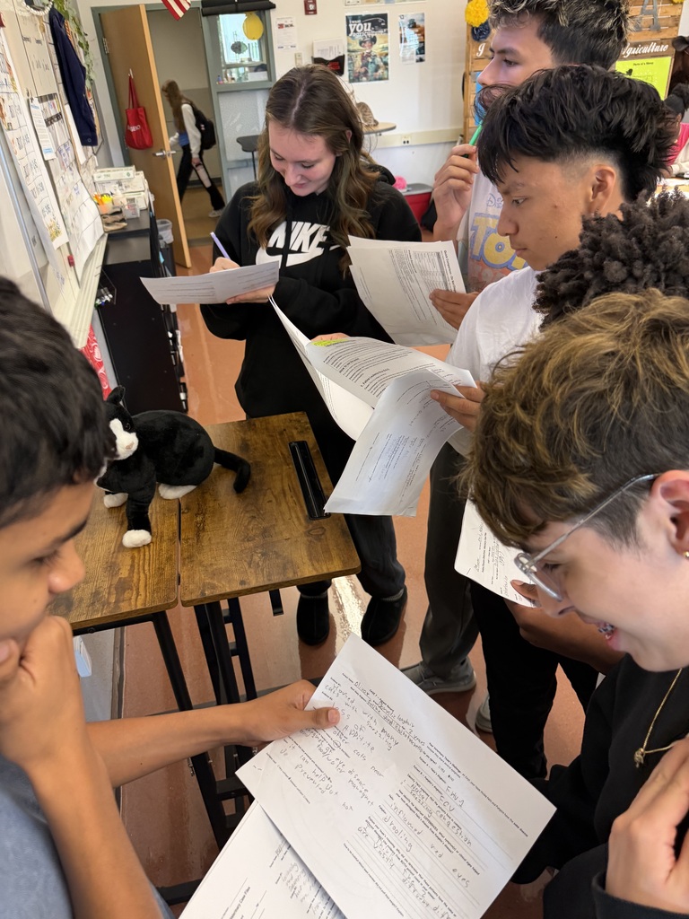Students in a classroom with papers, one holding a stuffed animal, others wearing glasses, some looking at papers.