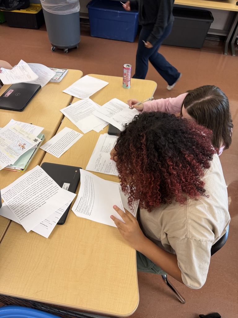 Two students sit at a table reviewing papers. One person leans over the table. Papers are scattered.