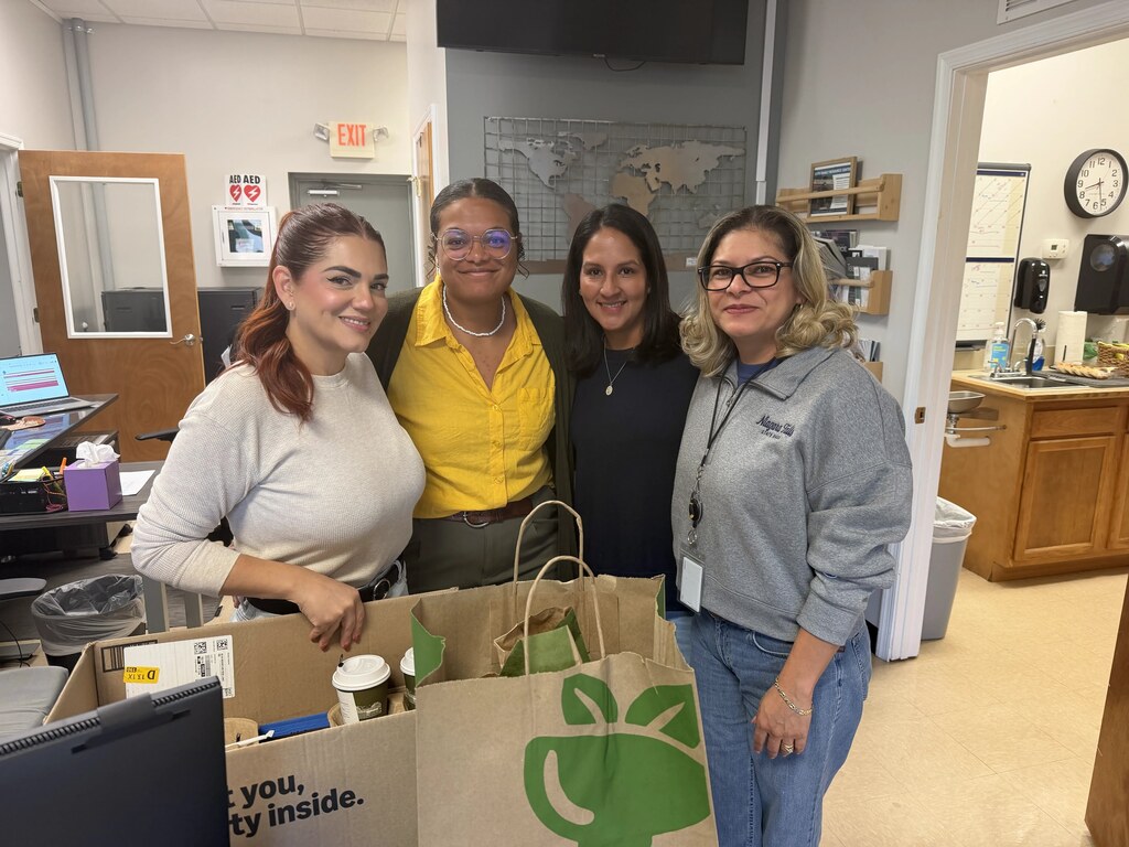 Four Family Resource Center staff members smile together in an office at Culpeper Middle School, standing behind boxes and bags of supplies and donations.