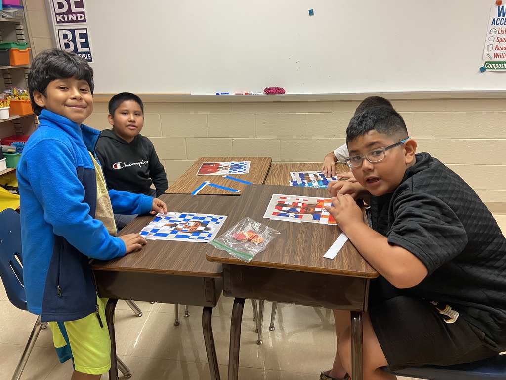 Three children sit around a table playing a game. Two wear glasses. A whiteboard is behind them.
