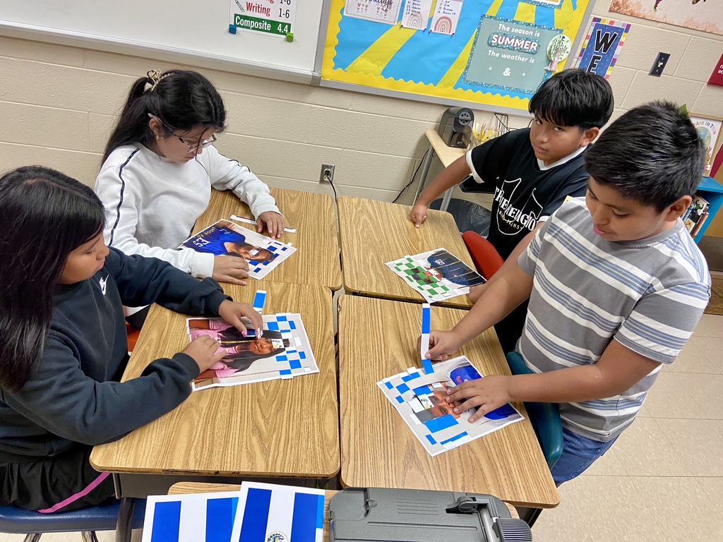 Four children at desks, engaged in an activity with papers and markers. One child holds a marker, pointing to a paper.