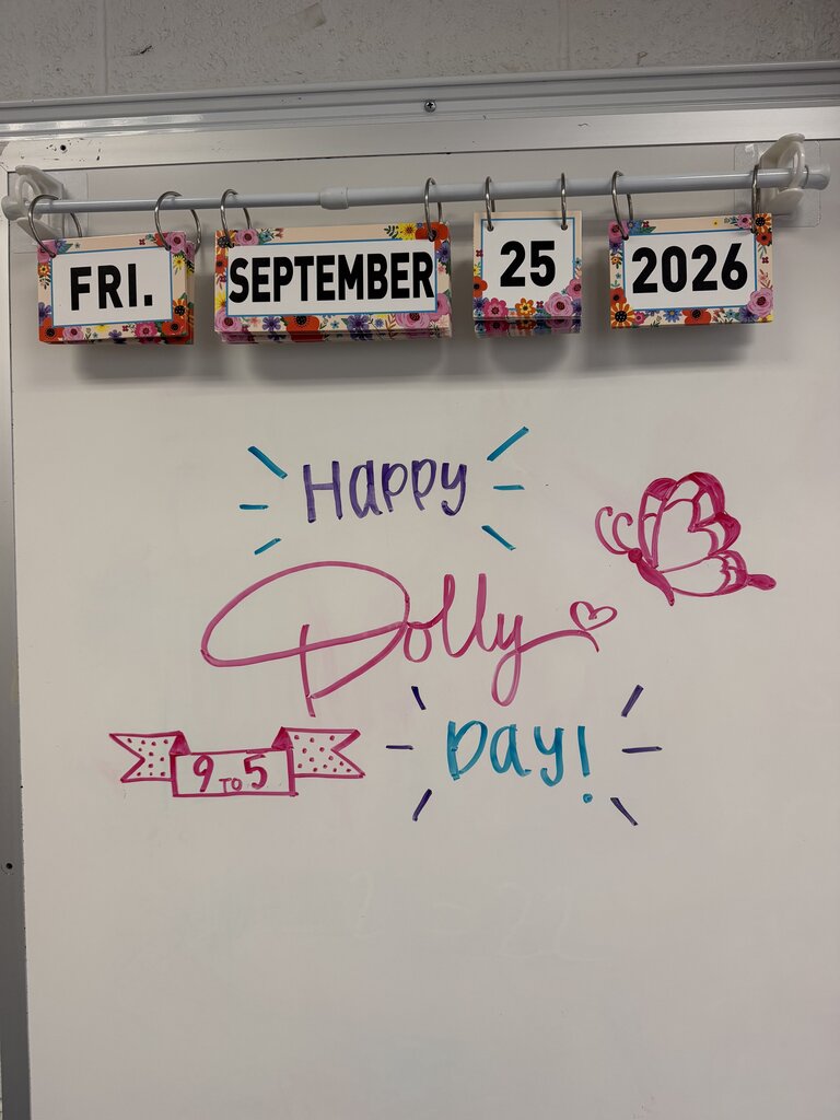Whiteboard with colorful labels displaying "Fri.", "September", "25", and "2026" above a message saying "Happy Dolly Day!" in pink, purple, and blue.