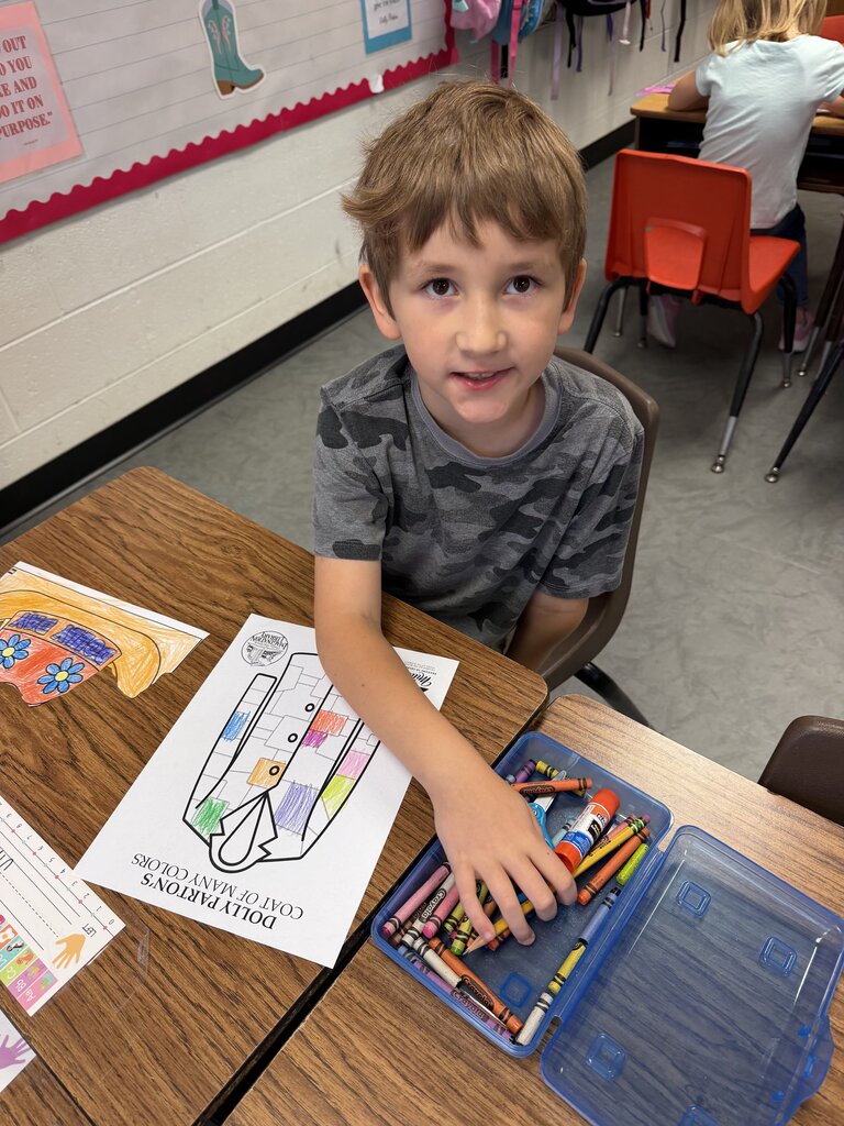 A young boy with brown hair uses crayons from a box at a desk. Behind him, another student sits at a desk.
