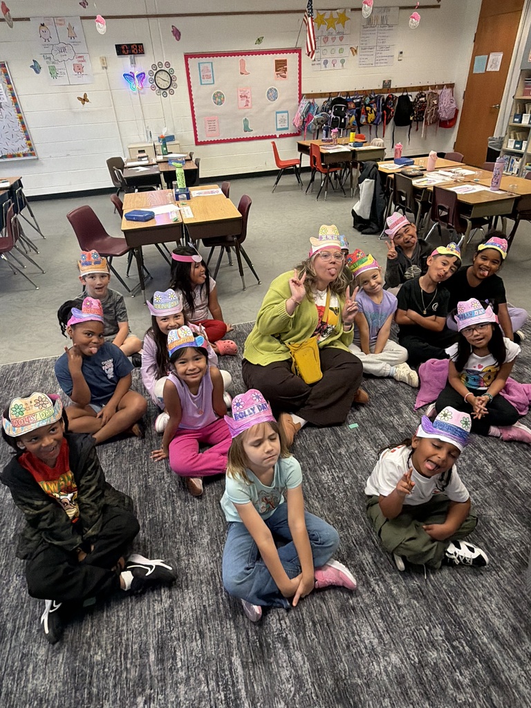 Kids and a woman sit on a carpeted floor in a classroom. Some wear hats.