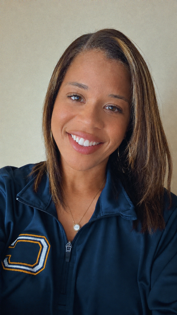 Woman with brown hair, wearing a navy blue sports jacket with gold and white letters. She smiles and poses.
