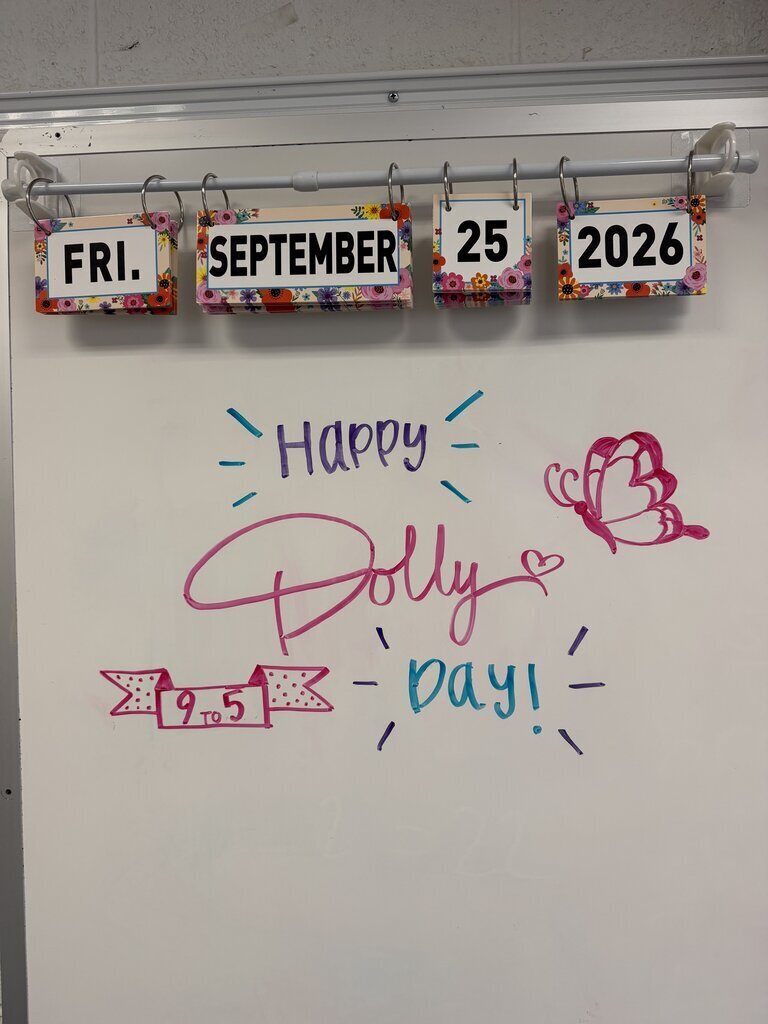 Whiteboard with colorful labels displaying "Fri.", "September", "25", and "2026" above a message saying "Happy Dolly Day!" in pink, purple, and blue.
