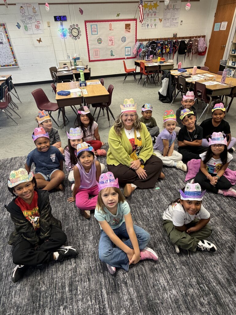 A group of children wearing party hats sits around a woman on a rug in a classroom.