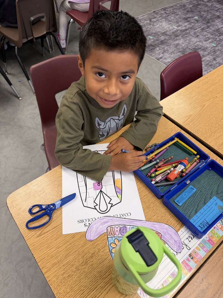 A boy sitting at a desk with a coloring book and scissors. Other school supplies are on the table.