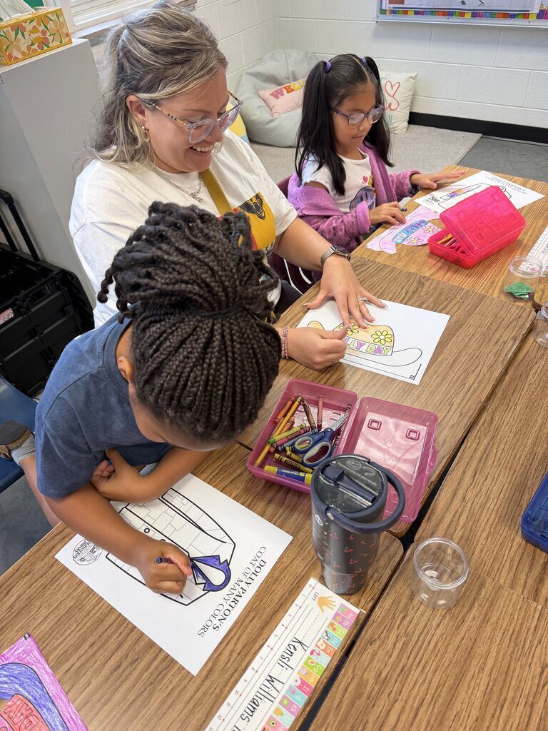 Three children draw on paper in a classroom. One wears glasses and smiles, while the others focus on their artwork.