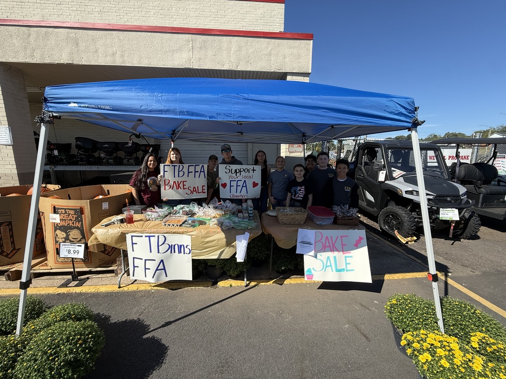 A group of people stand under a blue canopy, holding signs. Behind them is a building and parked vehicles.