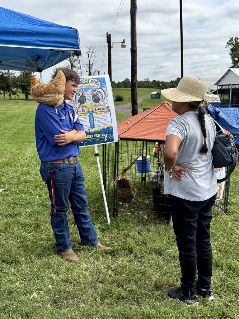 Two people converse by a tent, one with a poster and a chicken on his shoulder.