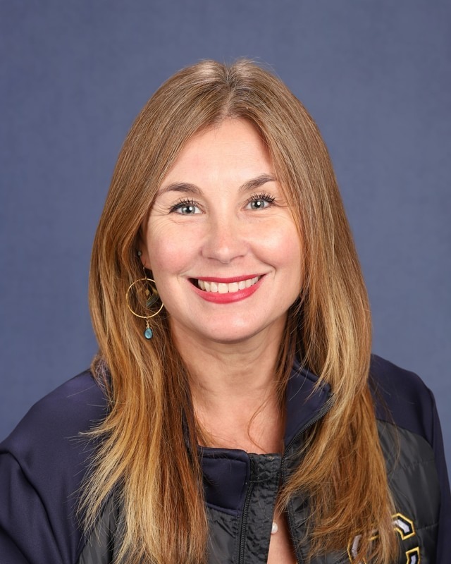 Woman with long blonde hair, smiling, wearing a navy blue jacket with a gold logo on the right side.