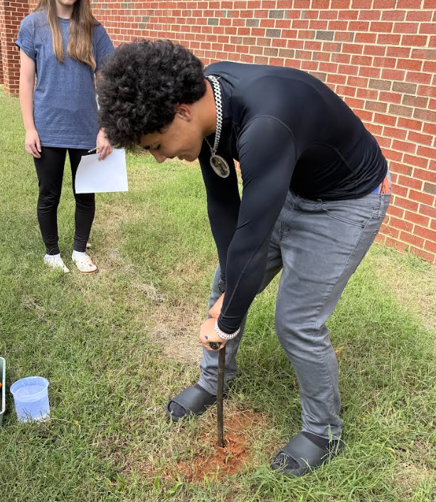 A person using a shovel on grass. A woman nearby holds a paper. Brick wall in background.