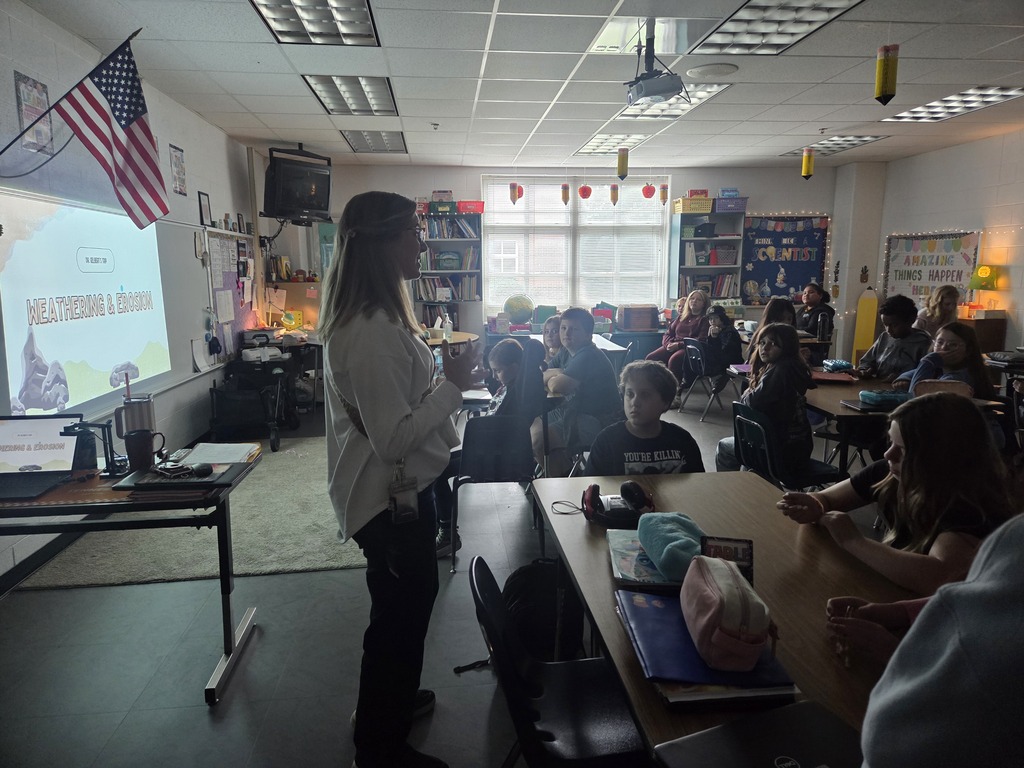 A woman stands before a classroom of seated students. A projector screen displays an image.