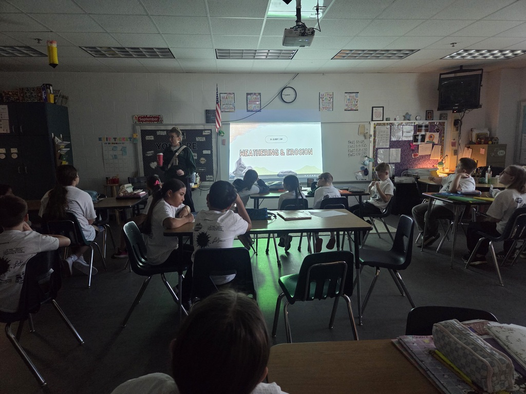 A classroom with students seated at tables. A teacher stands in front of a projector screen.