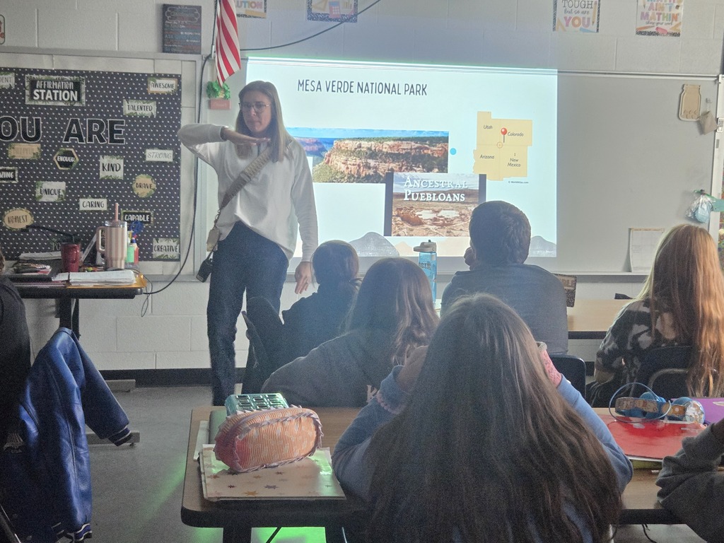 A woman teaching in a classroom with a projector screen displaying "Mesa Verde National Park" behind her.