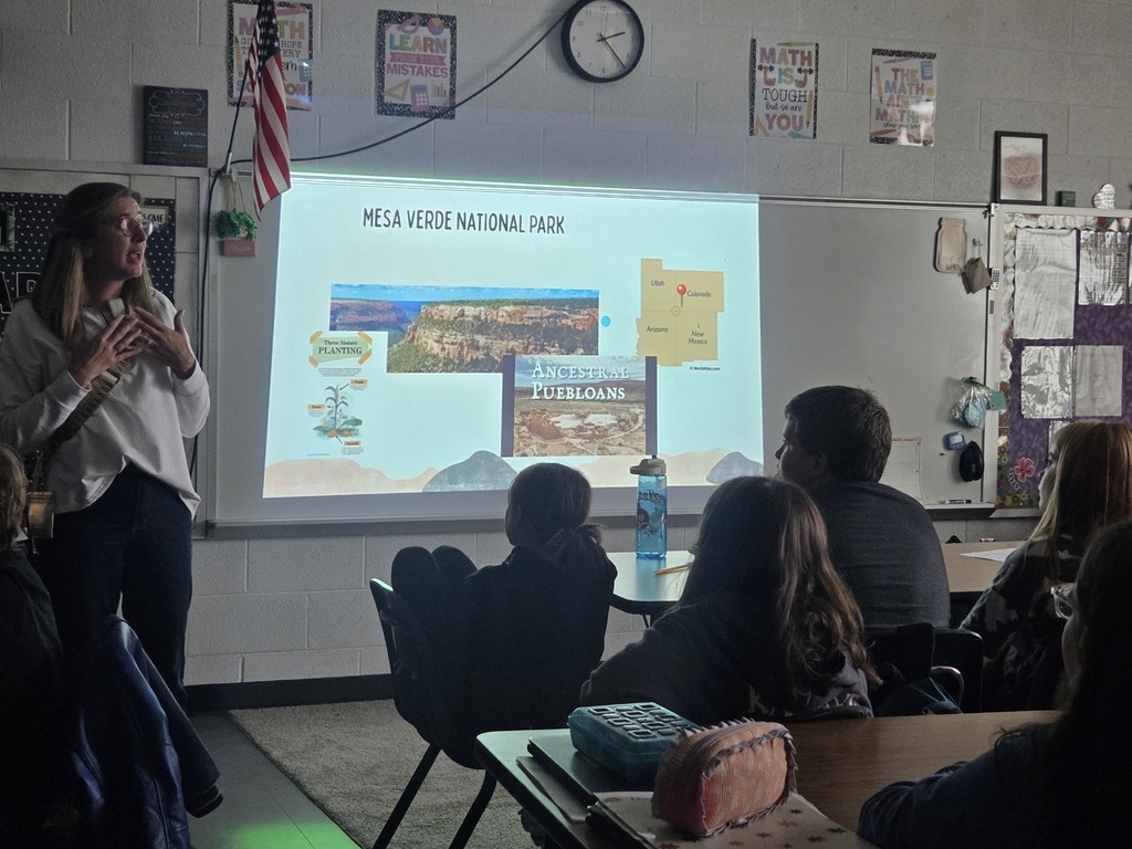 Woman standing before a projector screen while students sit in chairs. A clock and flags are on the wall.