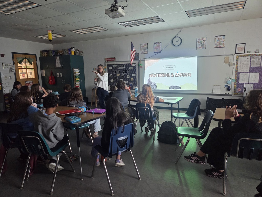 Classroom with students seated at tables; teacher standing by a projector screen displaying a presentation.