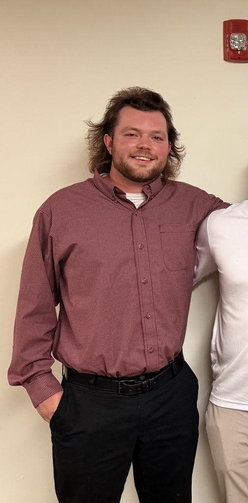 Man with long hair and beard wearing a maroon shirt and black pants standing next to another person.