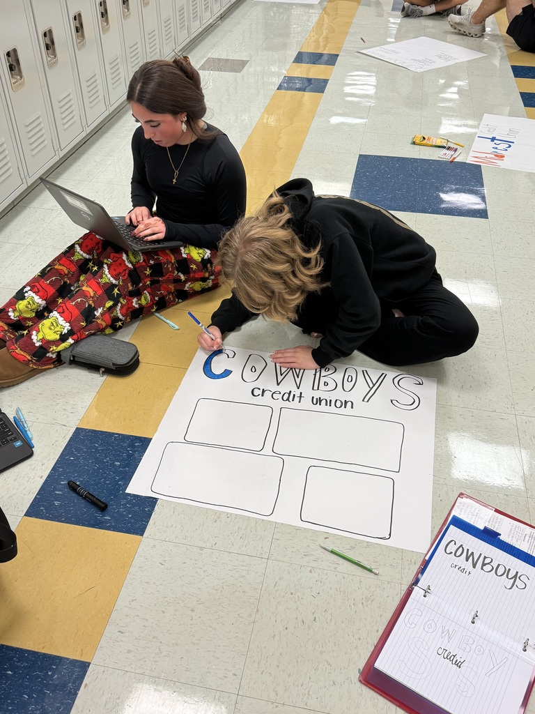 Two people in a school hallway with lockers, sitting on the floor. One is working on a laptop, and the other is writing on a large sheet of paper.