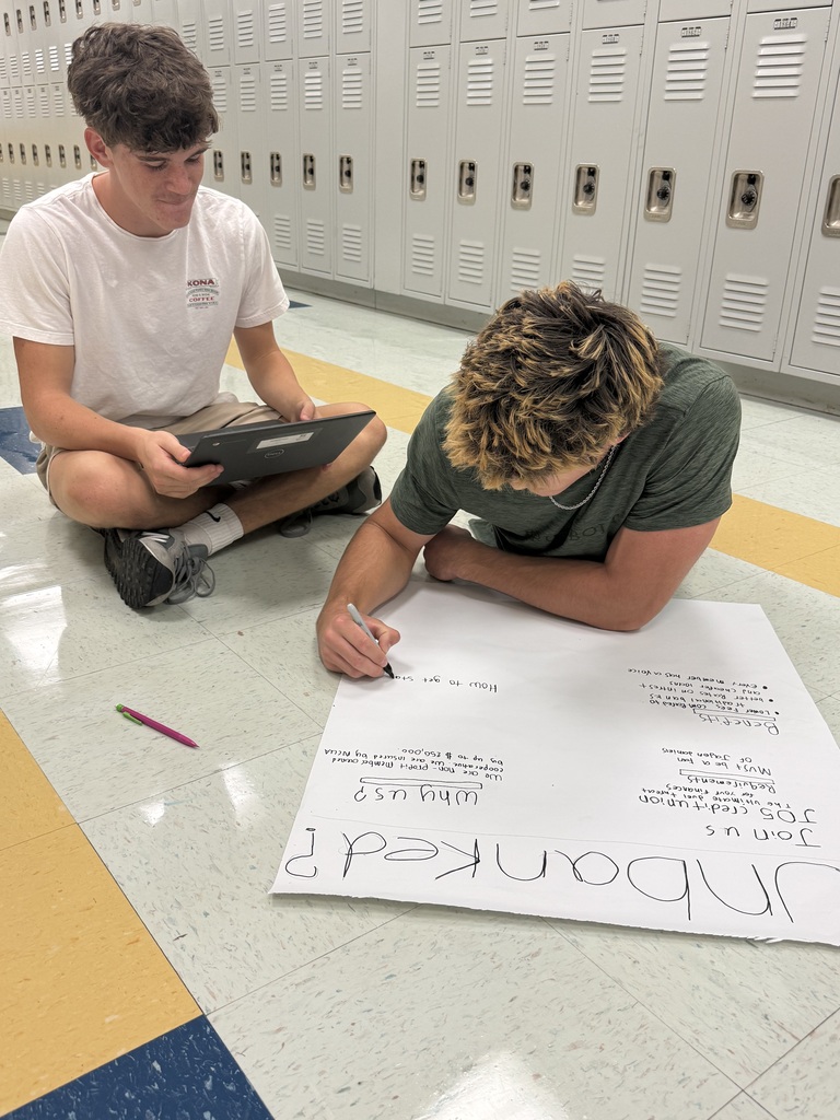 Two individuals seated on a floor, one writing on a large sheet of paper, with lockers in the background.