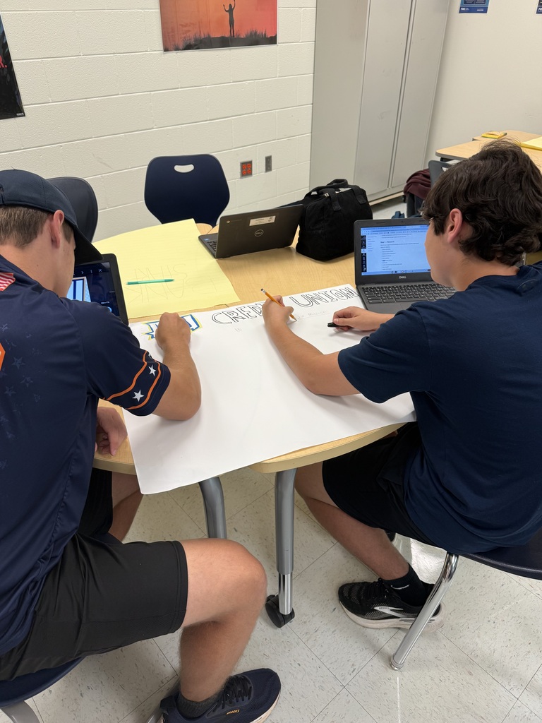 Two students sit at a table, facing each other, and each have a laptop open. They write on a piece of paper.
