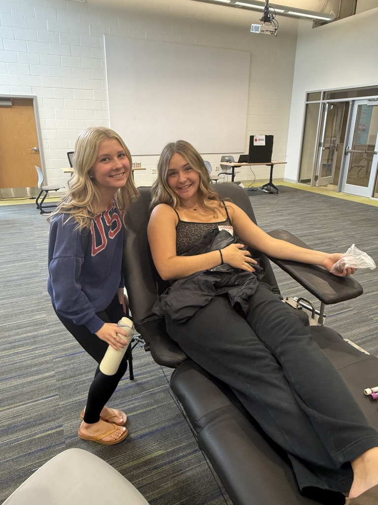 Two women are in a room. One sits in a chair with a bandage on her arm.
