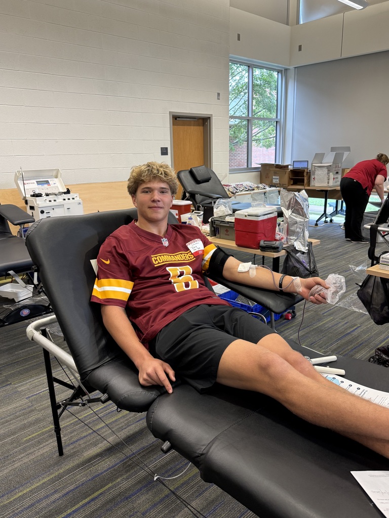 Man in red and black football jersey and shorts sits in a chair with a blood donation kit.
