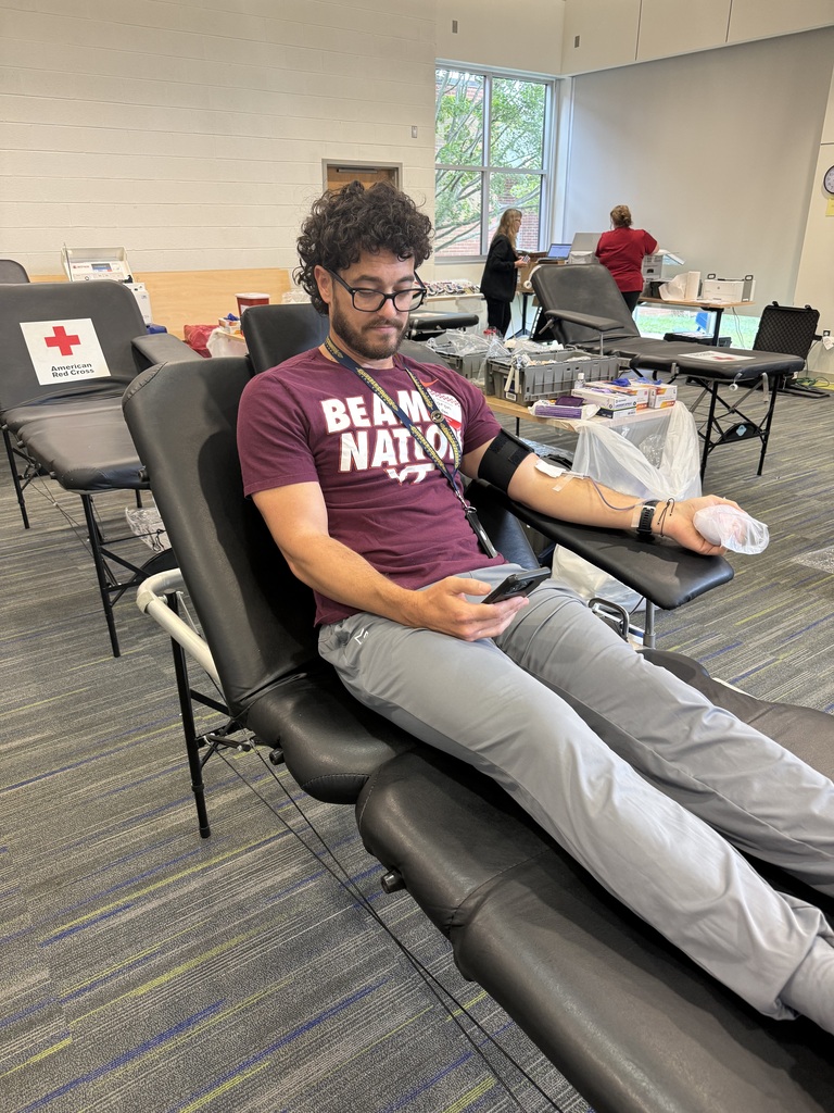 Man in maroon shirt with "Beam Nation" sits on reclining chair for blood donation with other chairs in background.