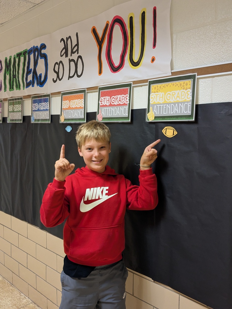 A boy in a red sweatshirt stands with fingers crossed in front of a bulletin board.