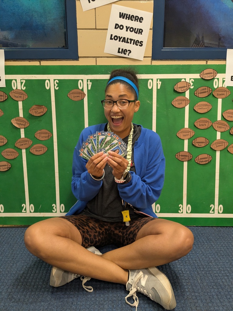 A person in a blue jacket and glasses sits on the floor, holding cards. They have a headband and are in front of a football-themed wall.