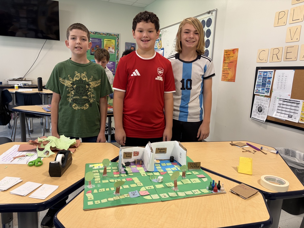 Three children at a table in a classroom with a map-like model in front of them.