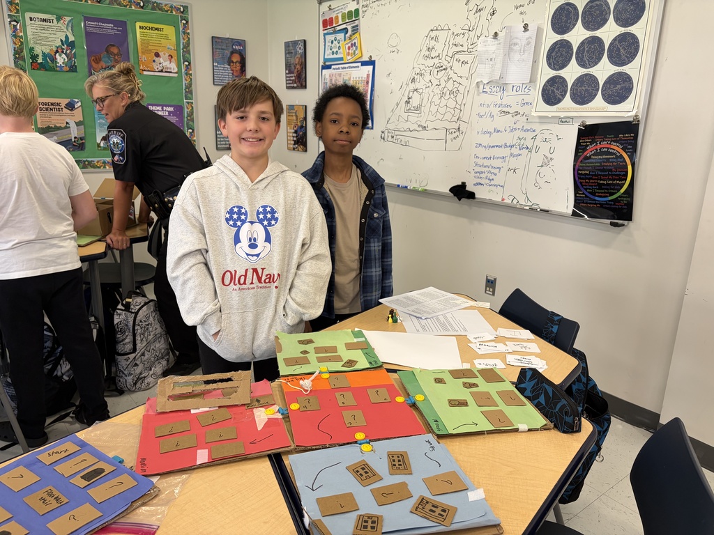 Two children stand by a table displaying various colorful, card-like constructions, with a whiteboard in the background.
