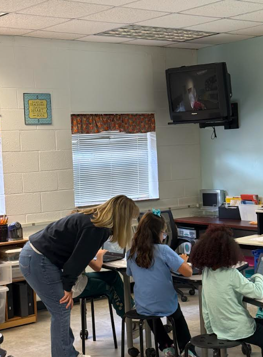 A teacher in a blue shirt leans over a student’s desk in a classroom with a mounted TV.
