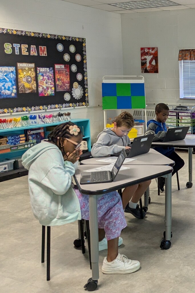 Four children seated around laptops on tables in a classroom. A bulletin board with STEAM and colorful posters adorns the wall.
