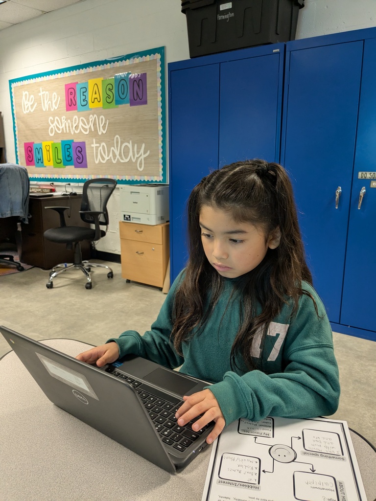 A young girl is sitting at a desk, focused on a laptop. Behind her, a motivational poster and blue storage cabinets.