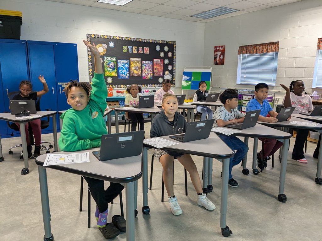 A classroom with students seated at desks, each using a laptop. Some students have their hands raised.