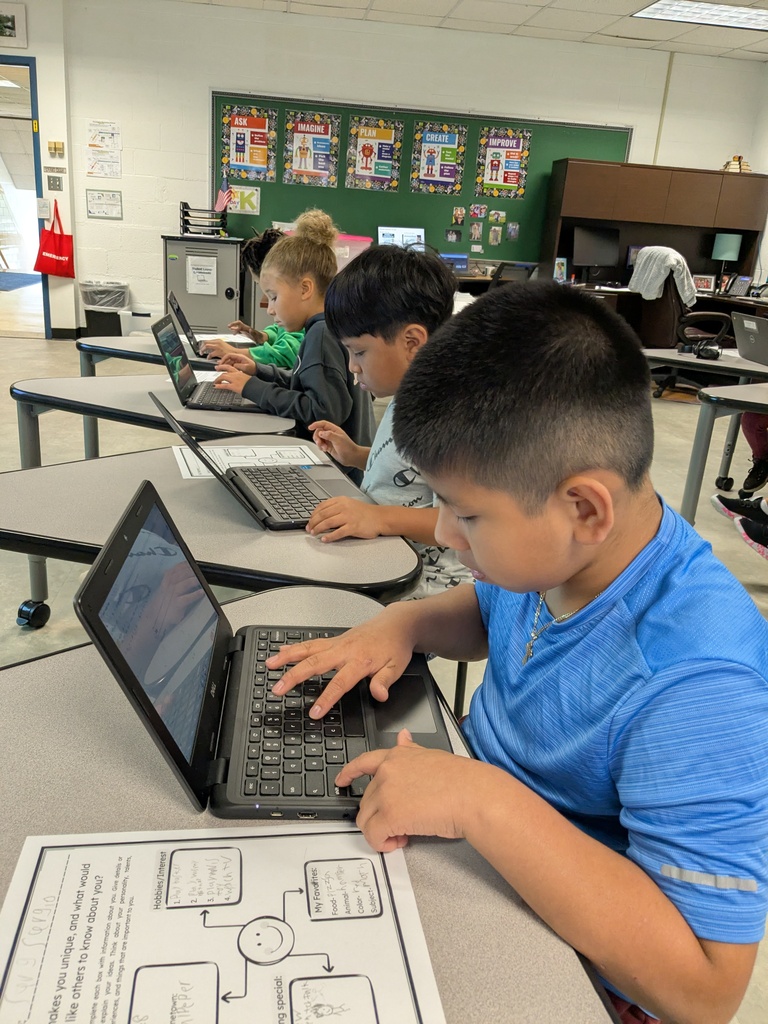 Four children sit at desks with laptops, engaged in typing. One student wears a blue shirt. Behind them are desks and a bulletin board.
