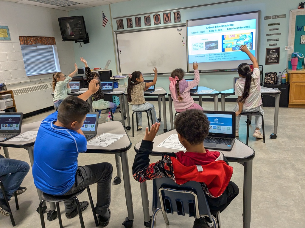 A classroom with children seated at desks, using laptops. Some are raising their hands. A projector screen on the wall.