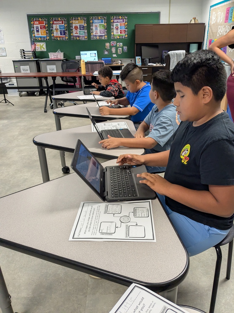 Four children are seated at desks in a classroom, each using a laptop. One child is wearing a black shirt.