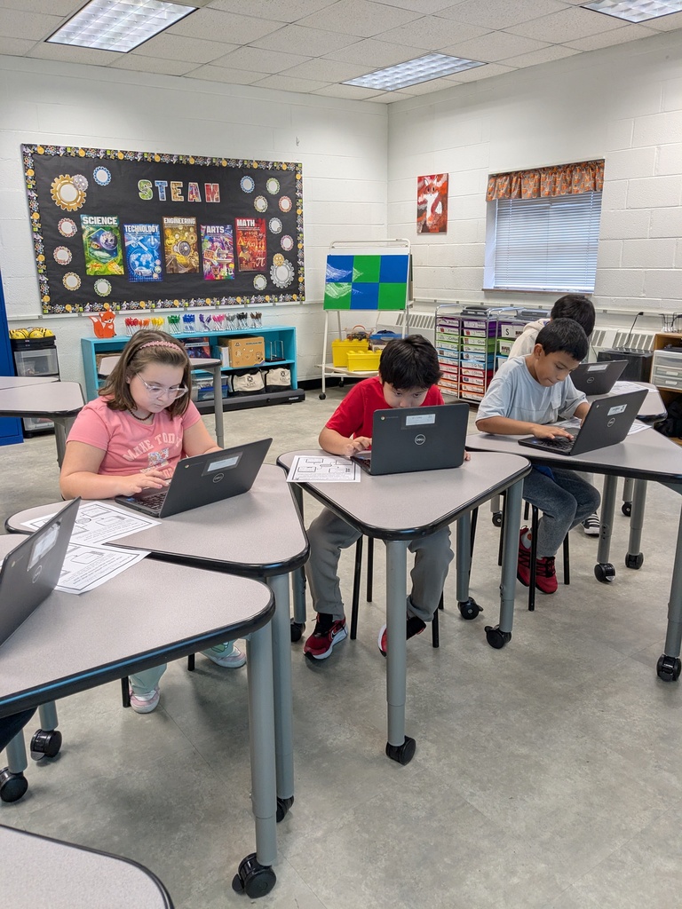 Four children in a classroom with tables and laptops. A bulletin board is on the wall.