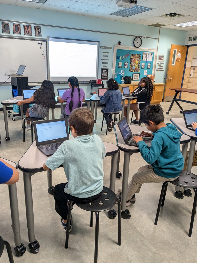 A classroom with children sitting on stools, using laptops. A whiteboard, projector screen, and clock are visible.